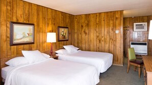 A hotel room with two beds, a wooden wall, a painting, a lamp, and a chair. - Sunset Motel (Moriarty)