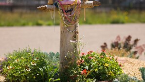A wooden sculpture with outstretched arms, adorned with colorful beads and chains, set in a garden with various plants and flowers. - Sunset Motel (Moriarty)