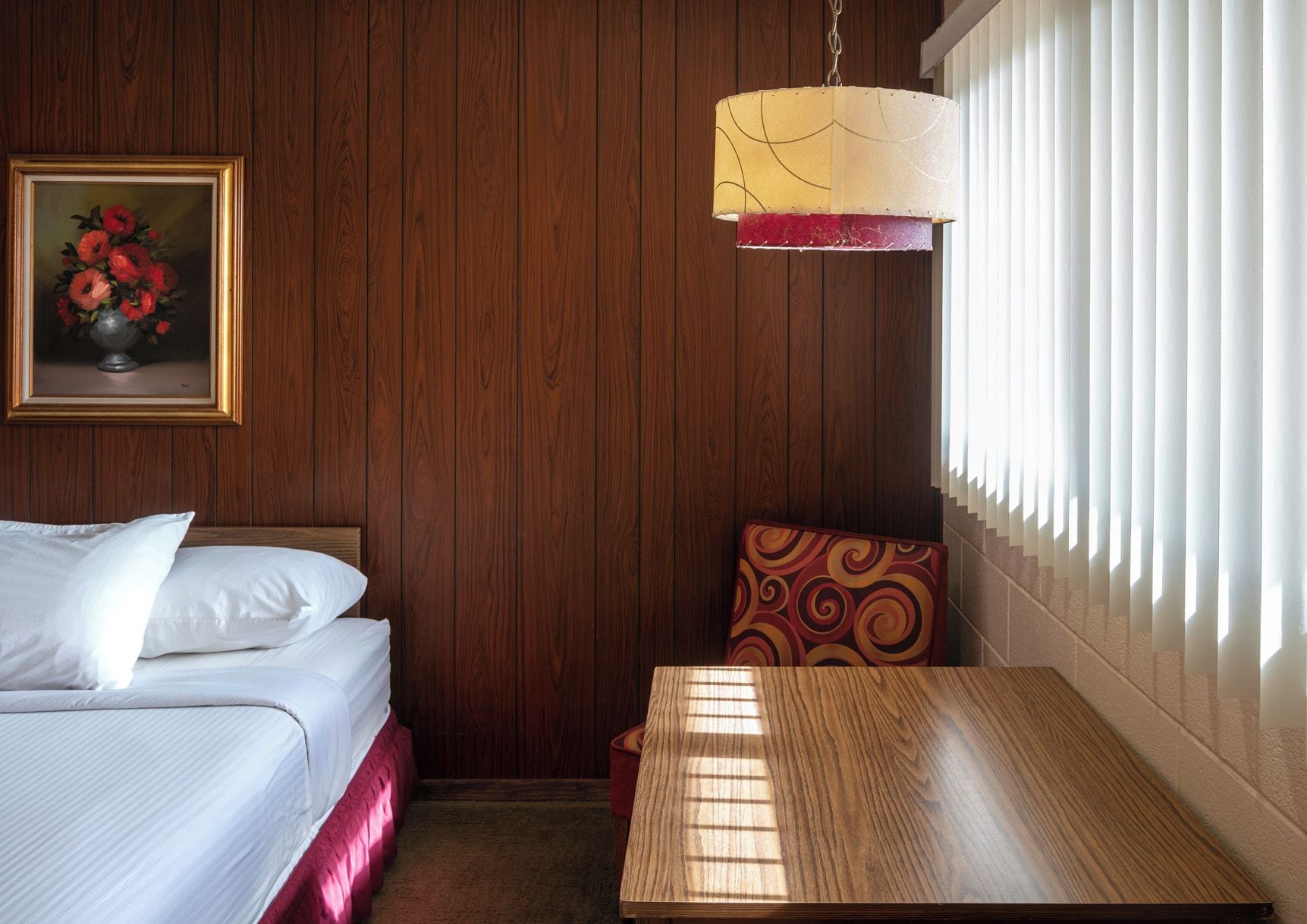 A hotel room with a wooden paneled wall, a bed with white linens, a table, a chair, a hanging lamp, and a window with blinds.