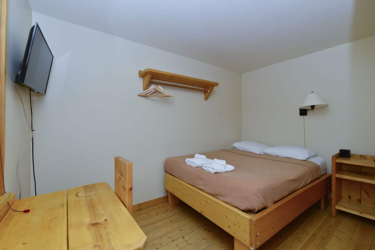 A single bed with a wooden frame, a bedside table, a wall-mounted TV, a hanging clothes rack, and a floor lamp.