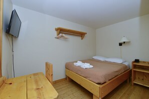 A single bed with a wooden frame, a bedside table, a wall-mounted TV, a hanging clothes rack, and a floor lamp.