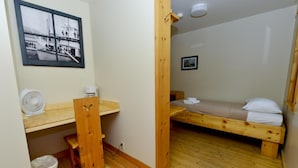 Una habitación pequeña y acogedora con una cama de madera, un escritorio y una silla. Hay un cuadro en la pared, un ventilador y un cubo de basura.