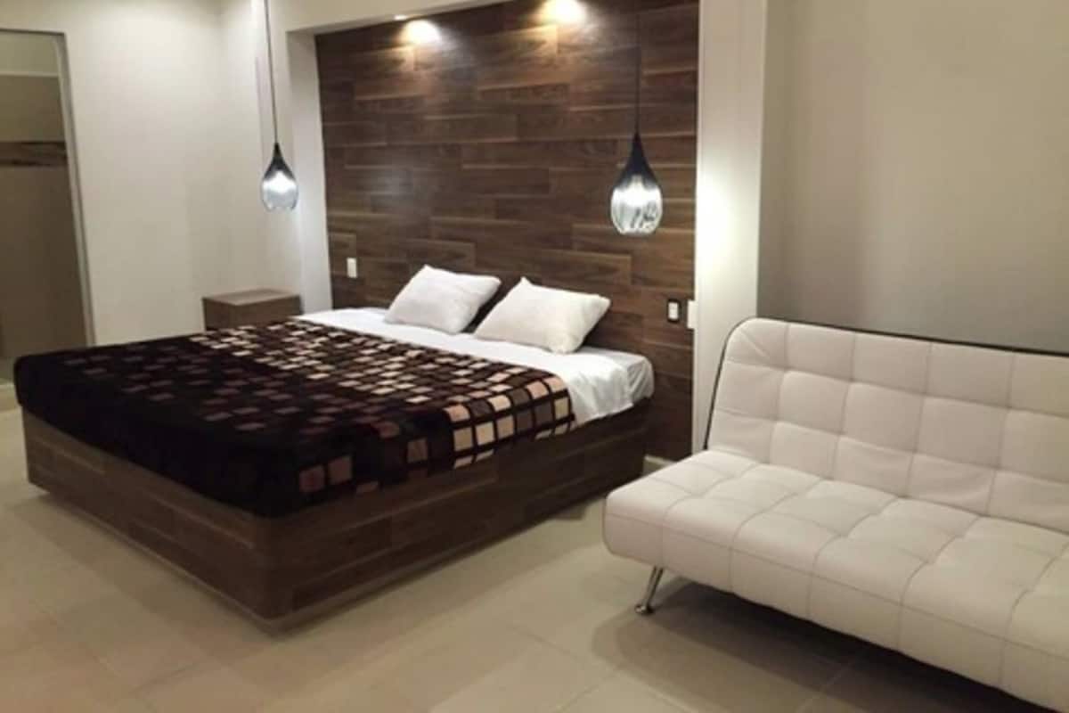Une chambre à coucher moderne avec une tête de lit en bois, un grand lit recouvert d’une couvre-lit à motifs, un canapé blanc et des luminaires suspendus.