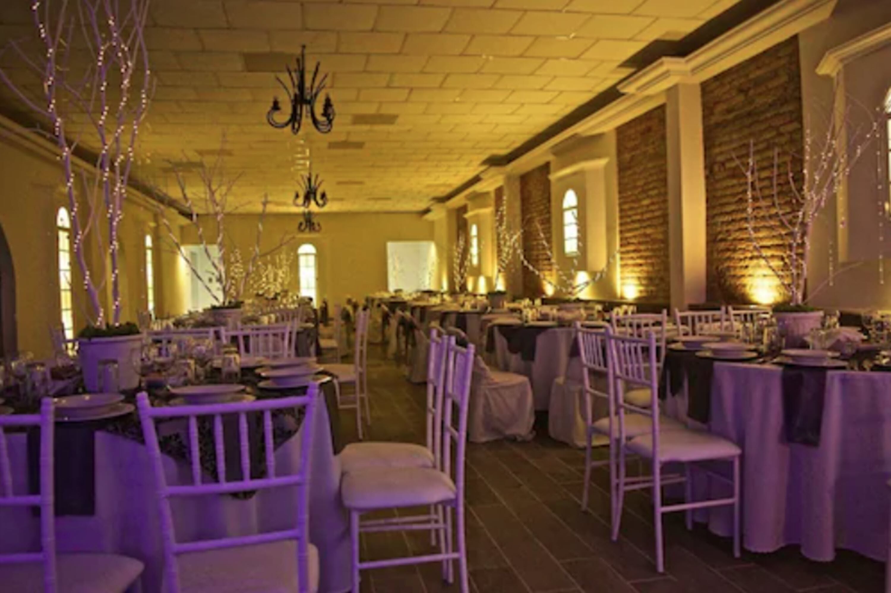 Un salón de banquetes con mesas dispuestas para un evento formal, con sillas blancas y mesas cubiertas con manteles.