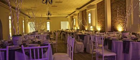 Un salón de banquetes con mesas dispuestas para un evento formal, con sillas blancas y mesas cubiertas con manteles.