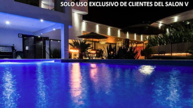 A nighttime view of a swimming pool with a patio area and outdoor seating, surrounded by modern architecture.