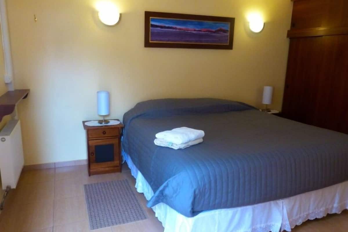 A neatly made bed with a blue bedspread and white pillows, a small bedside table with a lamp, and a painting on the wall.