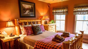 A cozy bedroom with a wooden headboard, a bed with checkered bedding, a bedside table with a lamp, and a window with curtains. - RoosterComb Inn (Keene Valley)