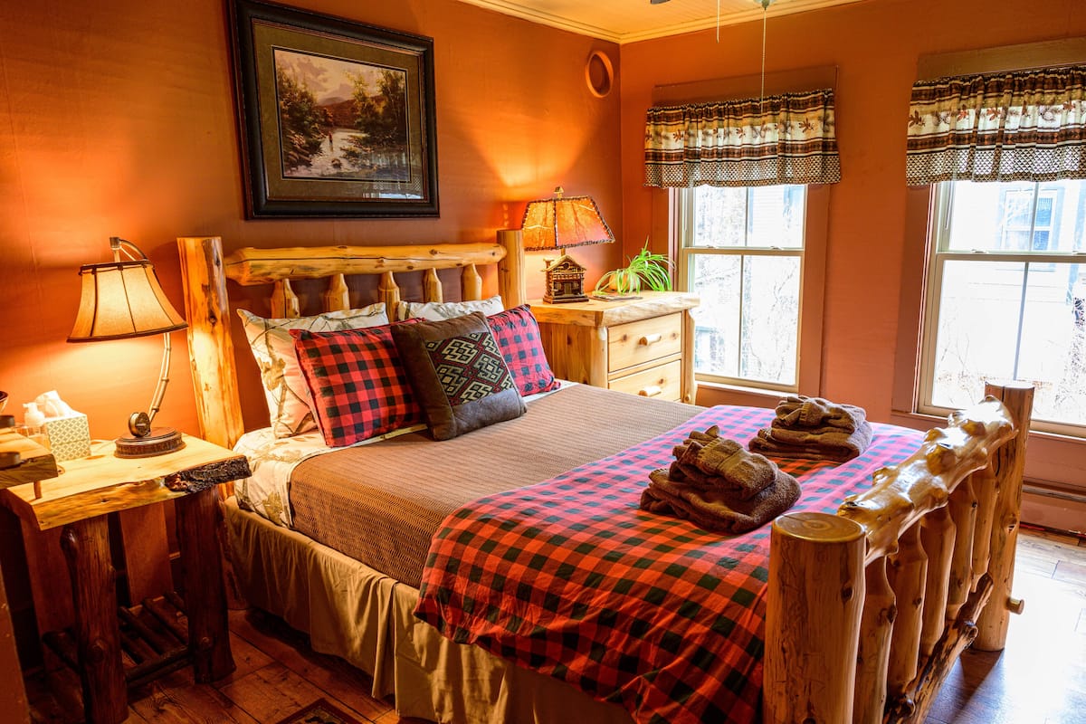 A cozy bedroom with a wooden headboard, a bed with checkered bedding, a bedside table with a lamp, and a window with curtains.