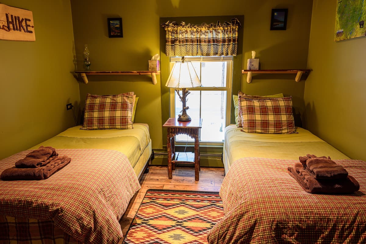 A room with two beds, each with a plaid bedspread and matching pillows, a small table with a lamp, and a window with curtains.