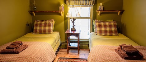 A room with two beds, each with a plaid bedspread and matching pillows, a small table with a lamp, and a window with curtains.