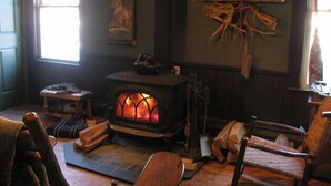A cozy room with a wood-burning stove, a chair, a small table, and a window. - RoosterComb Inn (Keene Valley)