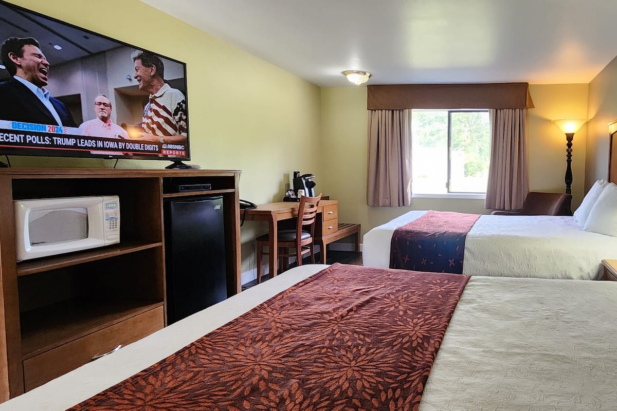 A hotel room with two beds, a desk, a TV, a microwave, and a window with curtains.