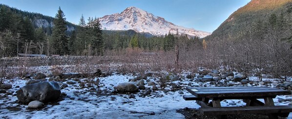 Paisaje nevado con una montaña al fondo, una zona rocosa con una mesa de picnic de madera.