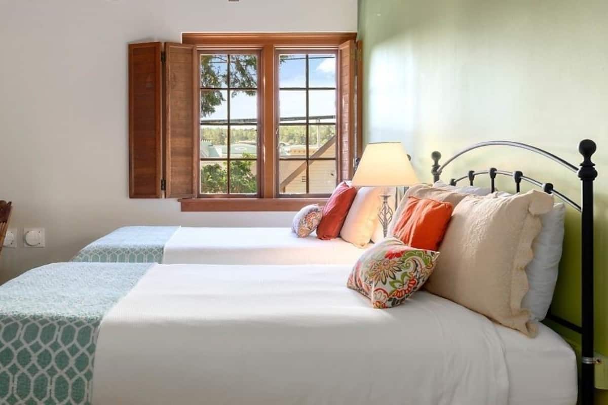 A bedroom with two beds, a window with open shutters, a lamp, and a chair.
