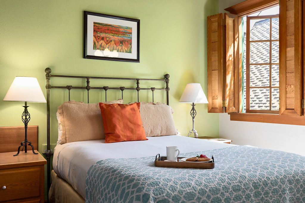 A neatly made bed with a tray of cookies and a cup, two bedside lamps, a framed picture on the wall, and a window with wooden shutters.