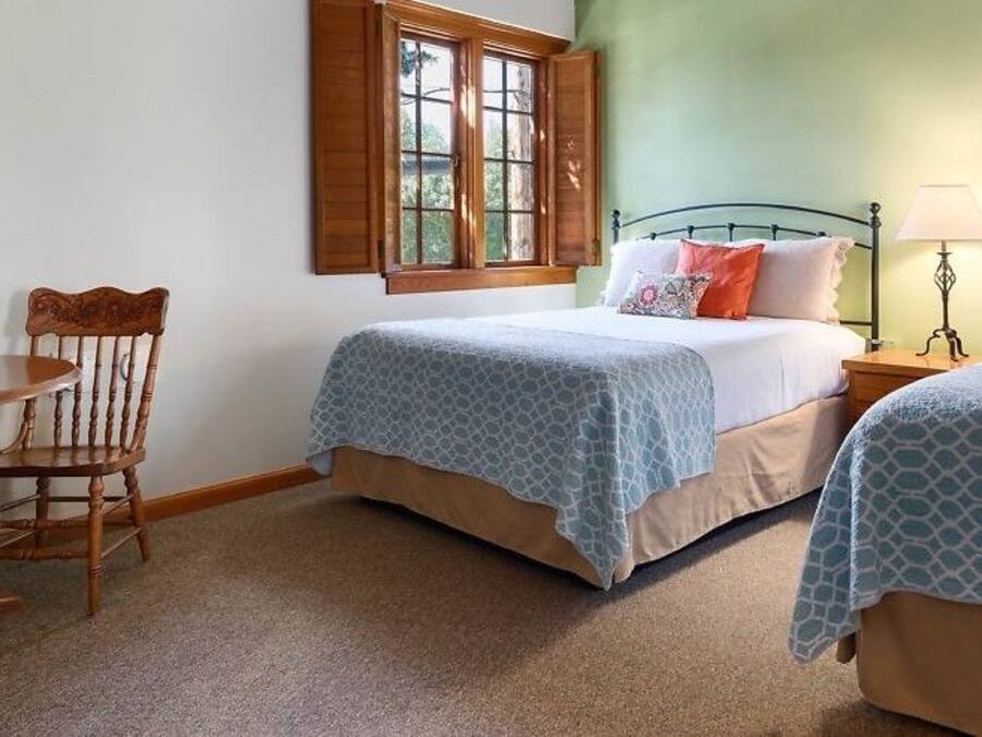 Un dormitorio con dos camas, una silla de madera, una mesita con un florero y una ventana con contraventanas.