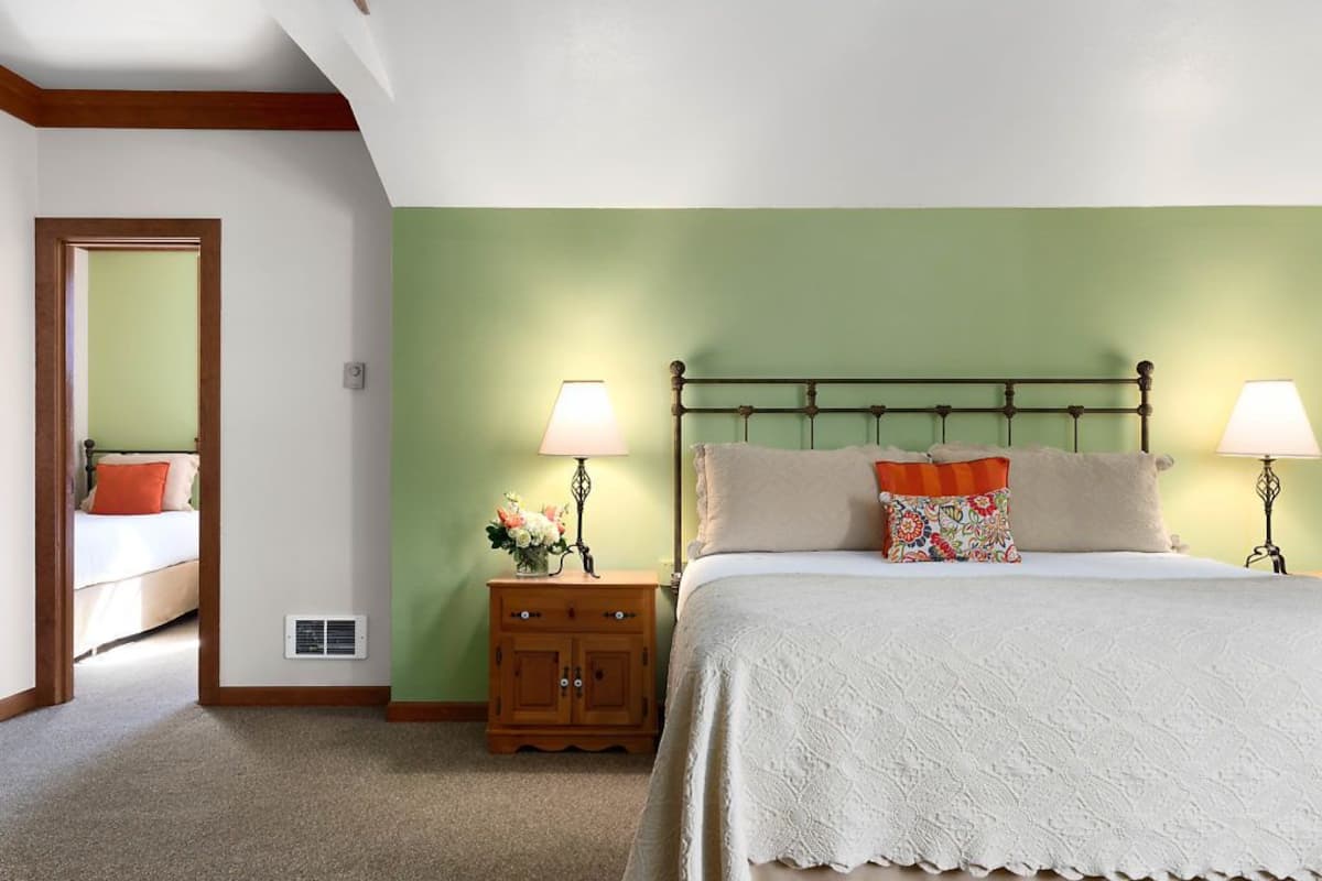 A bedroom with a bed, two bedside lamps, a nightstand with a vase of flowers, and a door leading to another room.