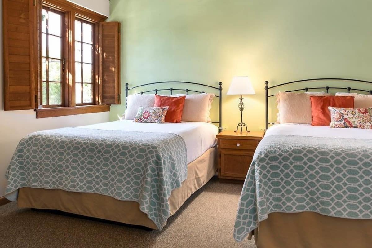 A room with two beds, each with a patterned bedspread, a nightstand with a lamp, and a window with wooden shutters.