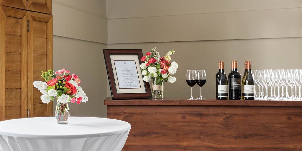 A wooden reception desk with a framed certificate, a vase of flowers, wine bottles, and glasses.
