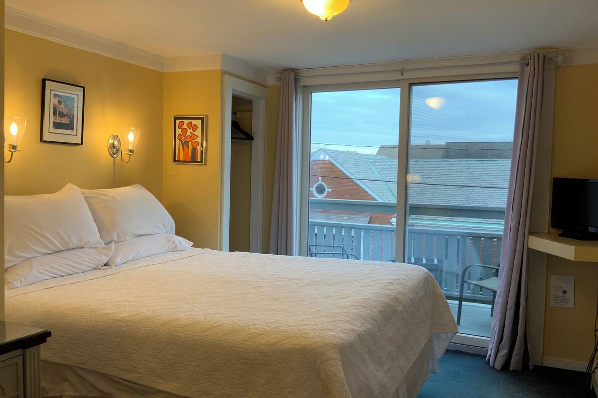 A hotel room with a large bed, a television on a stand, a balcony with a view of buildings, and a ceiling light.