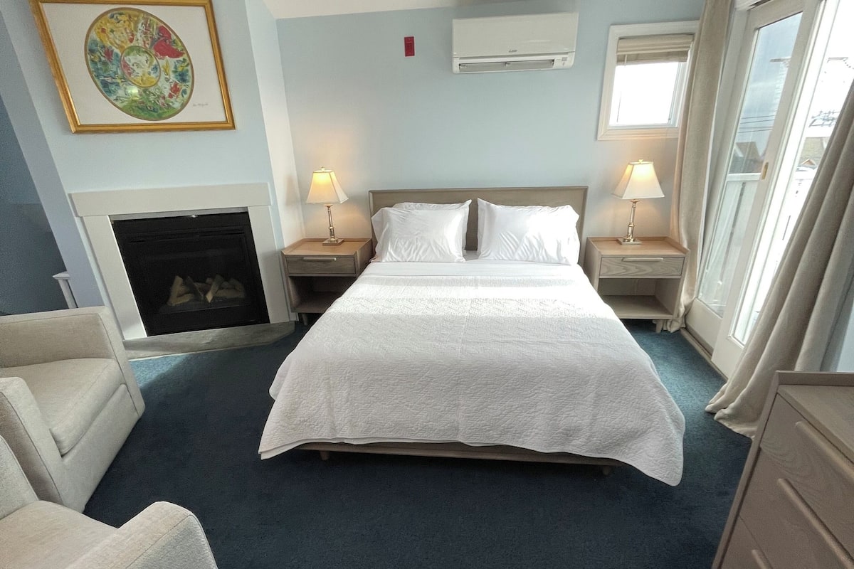 A hotel room with a large bed, two bedside tables with lamps, a fireplace, and a large window with curtains.