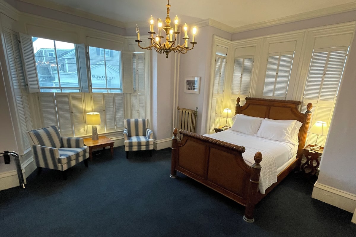 A bedroom with a large bed, two armchairs, a chandelier, and a view of the street through the windows.