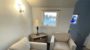 Une petite pièce accueillante, avec deux fauteuils beiges, une petite table surmontée d’une lampe, une fenêtre donnant sur les arbres et un petit vitrail.