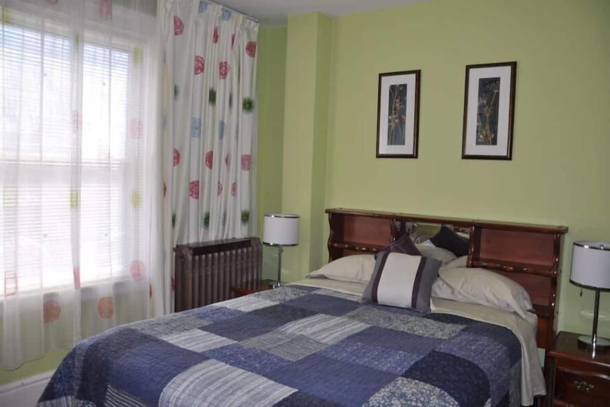 A bedroom with a large bed, two framed pictures on the wall, a radiator, and a window with curtains.