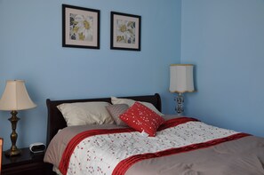 Ein ordentlich gemachtes Bett mit einer rot-weißen Steude, zwei gerahmte Blumenbilder an der Wand und Nachttischlampen.