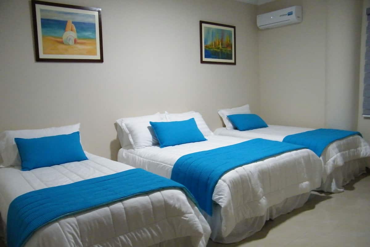 A hotel room with three beds, each with white bedding and blue pillows. There are two framed artworks on the walls and an air conditioning unit mounted on the wall.