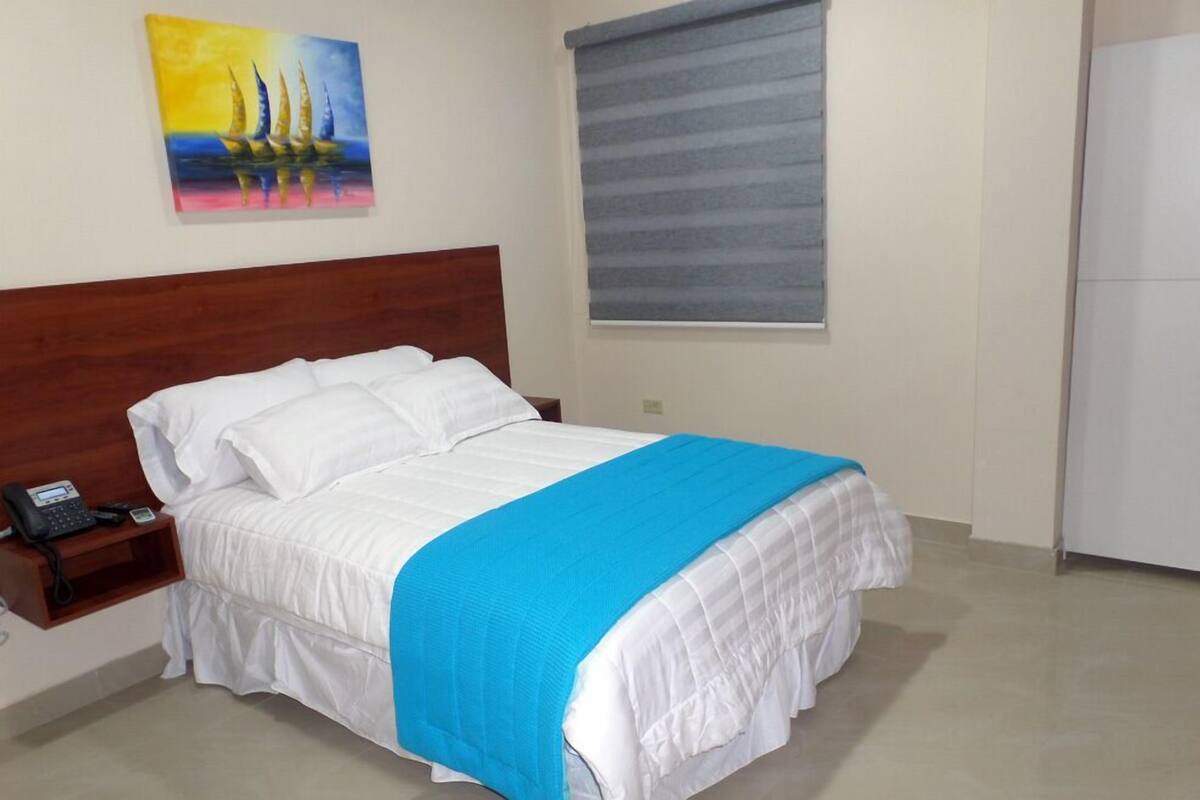 A neatly made bed with a blue blanket, a bedside phone, and a painting on the wall.