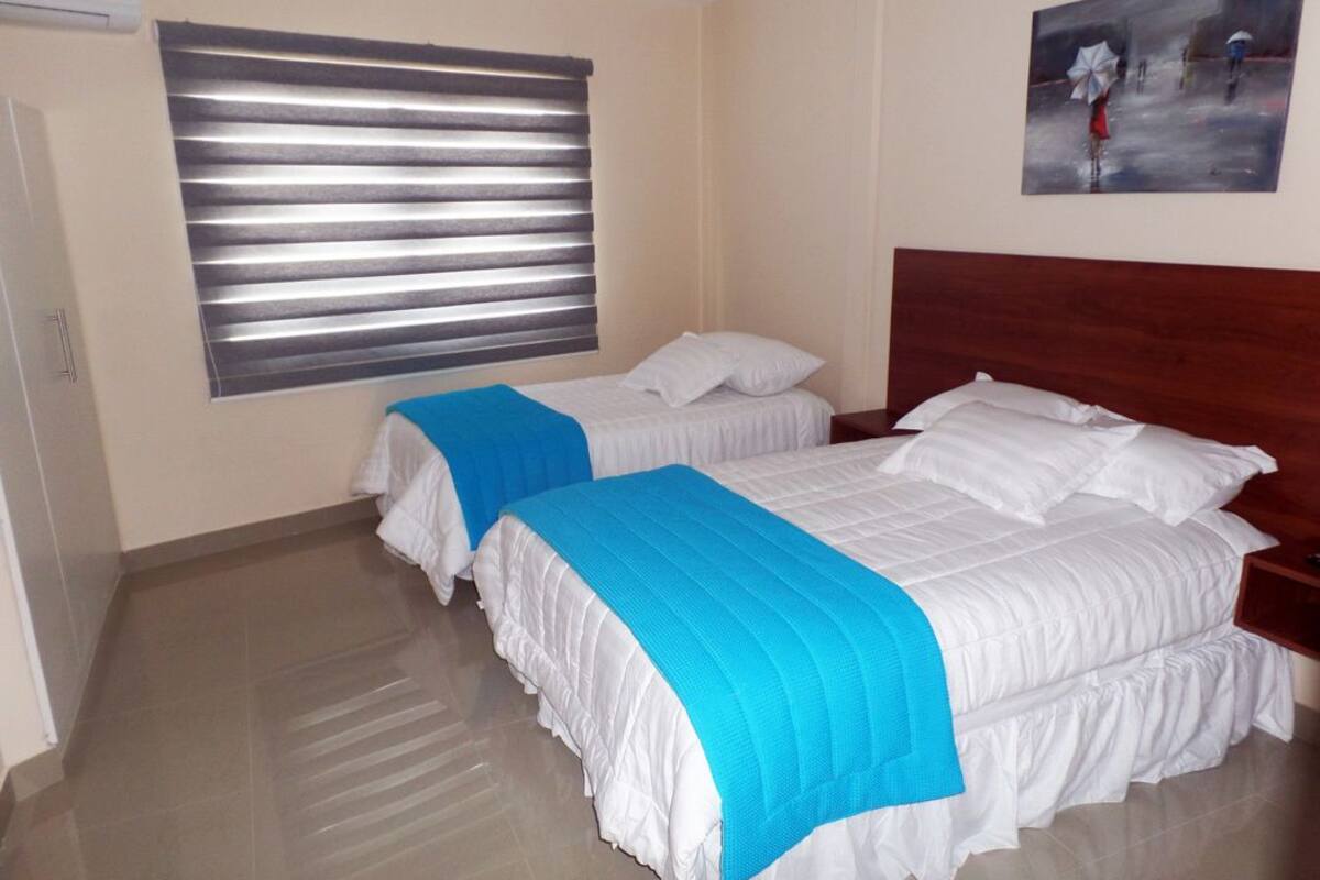 A hotel room with two beds, a window with blinds, a painting on the wall, and a ceiling-mounted air conditioner.