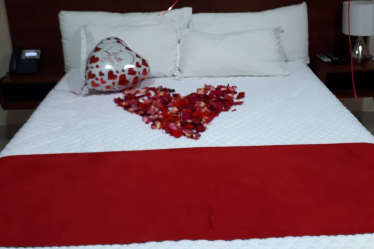 A neatly made bed with a red blanket, white pillows, and a heart-shaped arrangement of rose petals.