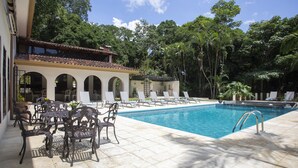 A swimming pool with lounge chairs and tables, surrounded by trees and a building with arches. - Villa San Ignacio (Tambor)