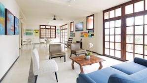 A spacious living area with a blue sofa, a wooden coffee table, and a dining table with chairs. There are large windows with wooden frames and artwork on the walls. - Villa San Ignacio (Tambor)