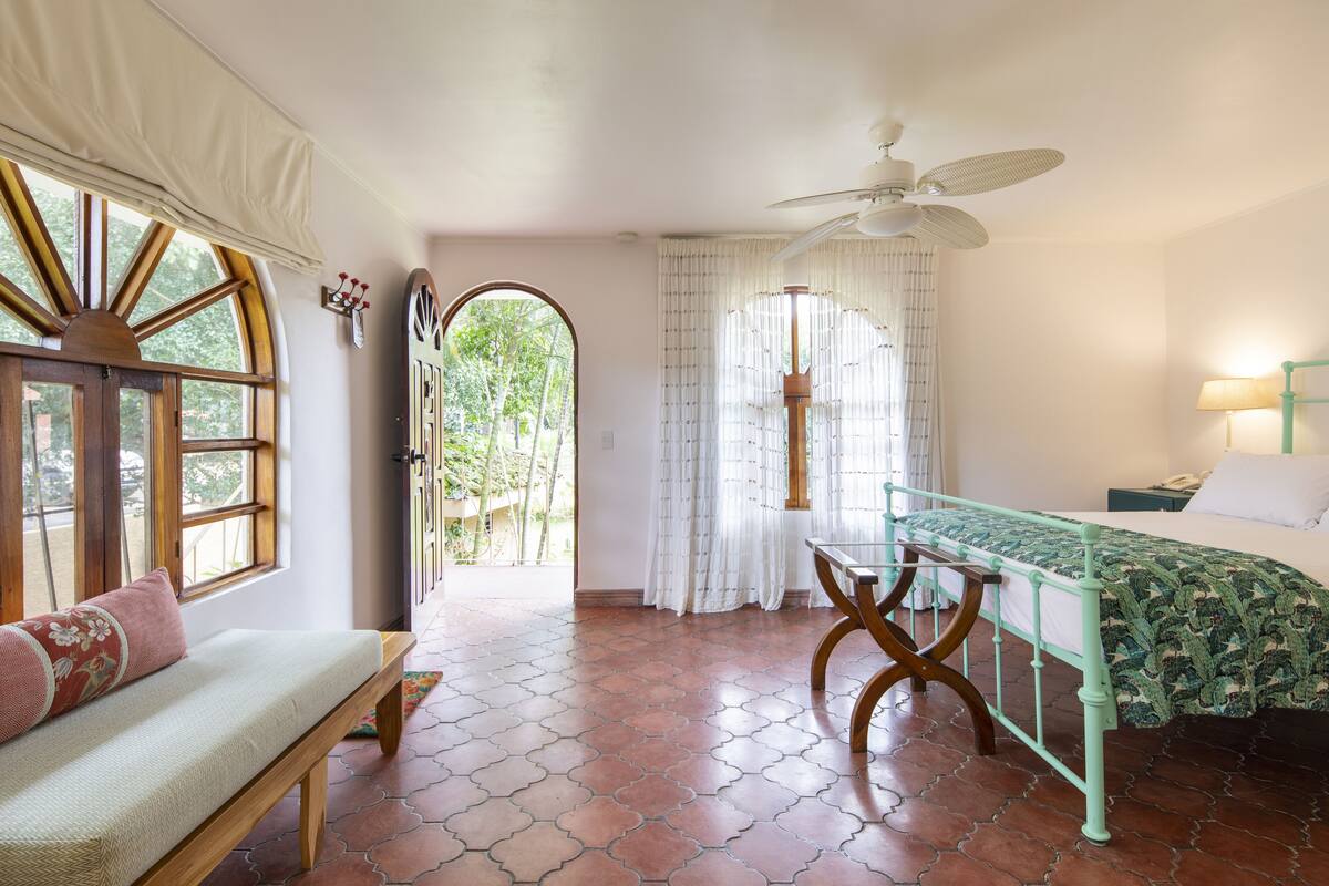 A room with a bed, a bench, a table, and a ceiling fan.