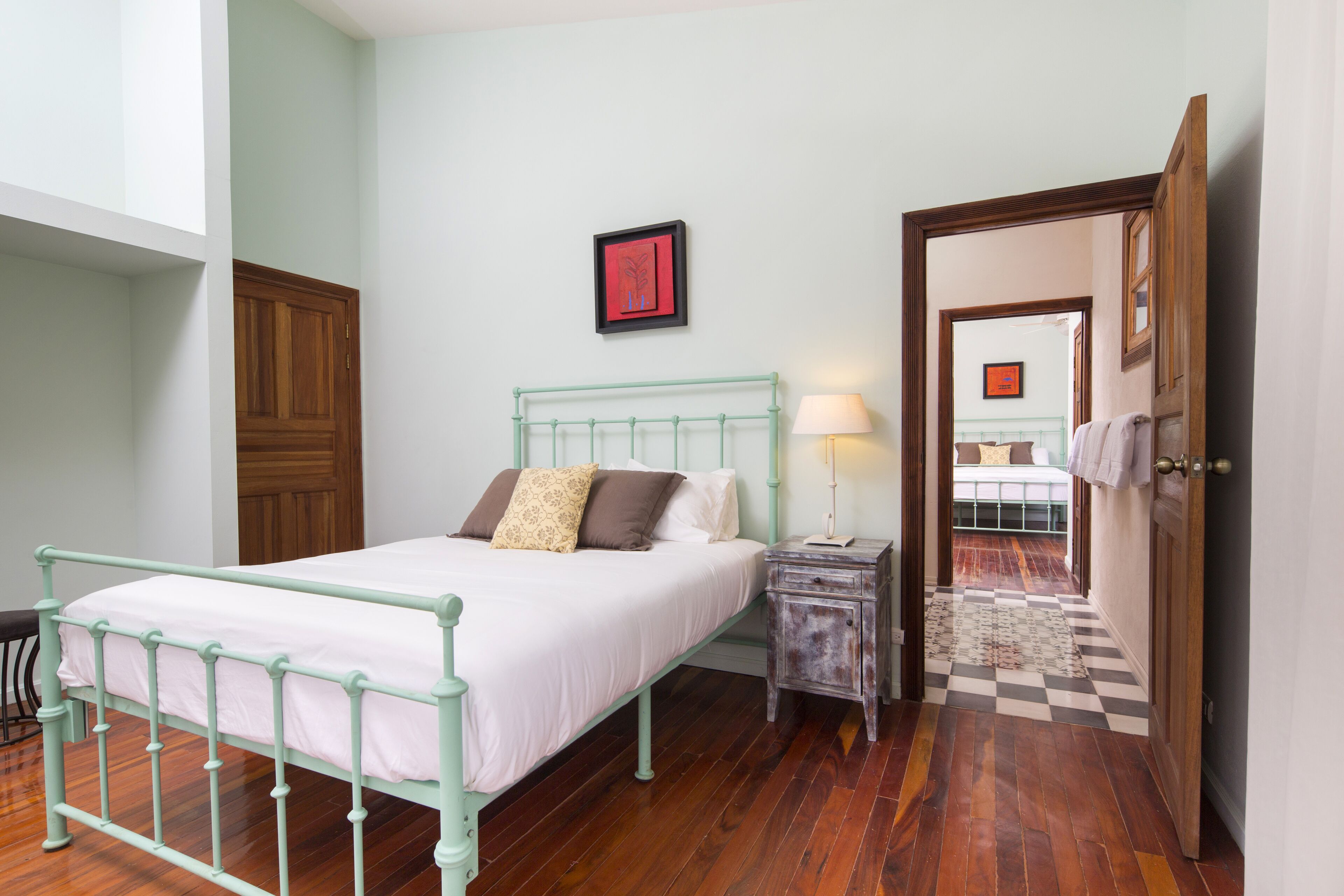 A bedroom with a bed, bedside table, lamp, and a door leading to another room.