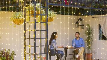 A couple seated at a table with string lights overhead, surrounded by potted plants and a pergola structure.