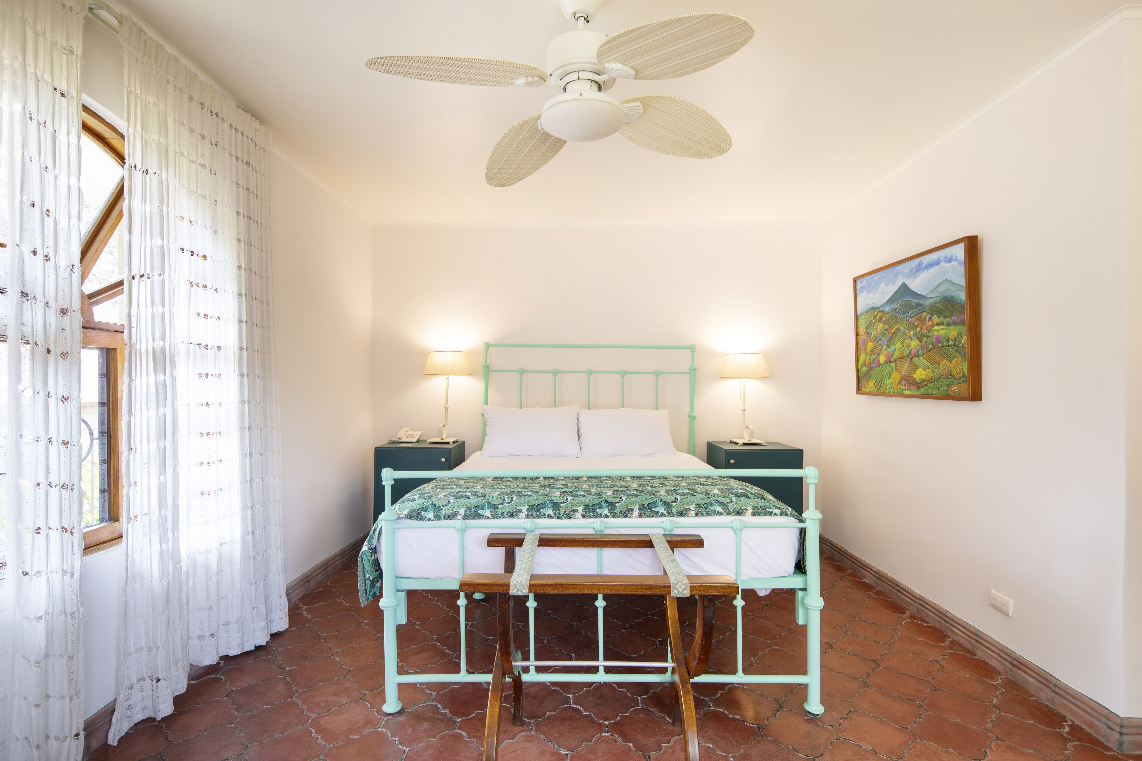 A bedroom with a bed, a ceiling fan, a window with curtains, a nightstand, and a wall-mounted painting.