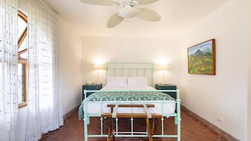 Quarto com cama, ventoinha no teto, janela com cortinas, mesa de cabeceira e um quadro pendurado na parede.