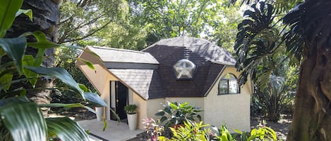 A small house with a sloped roof, surrounded by lush greenery and a variety of plants.