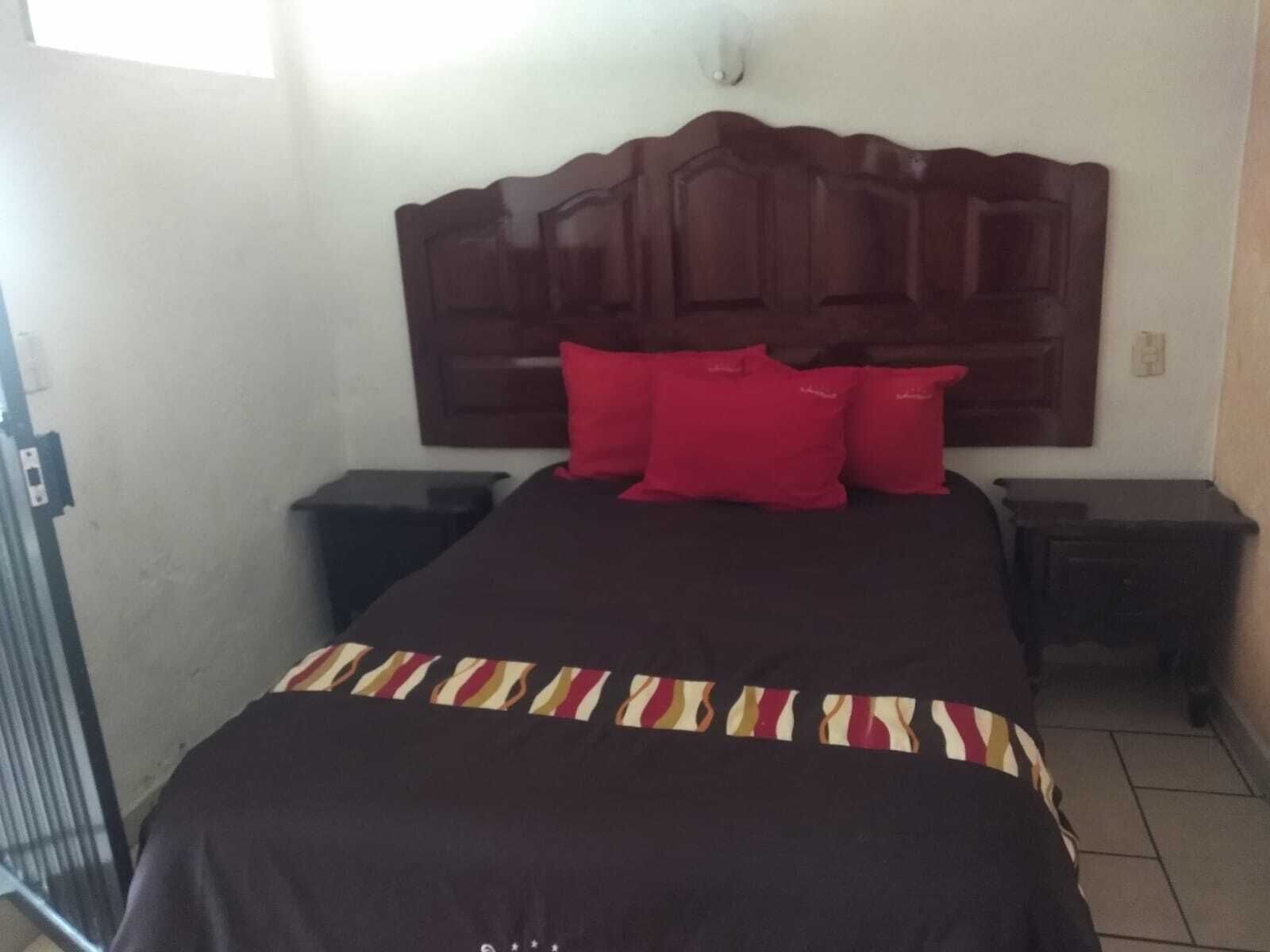 Habitación de hotel con una cama grande, un cabecero de madera, dos mesitas de noche y almohadas rojas.