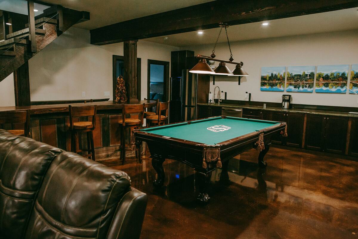 Mesa de bilhar em um espaço com piso de madeira, área de bar e escada.
