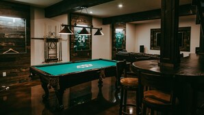 Une table de billard recouverte d’un drap vert, entourée de chaises en bois et d’un coin bar avec des tabourets.