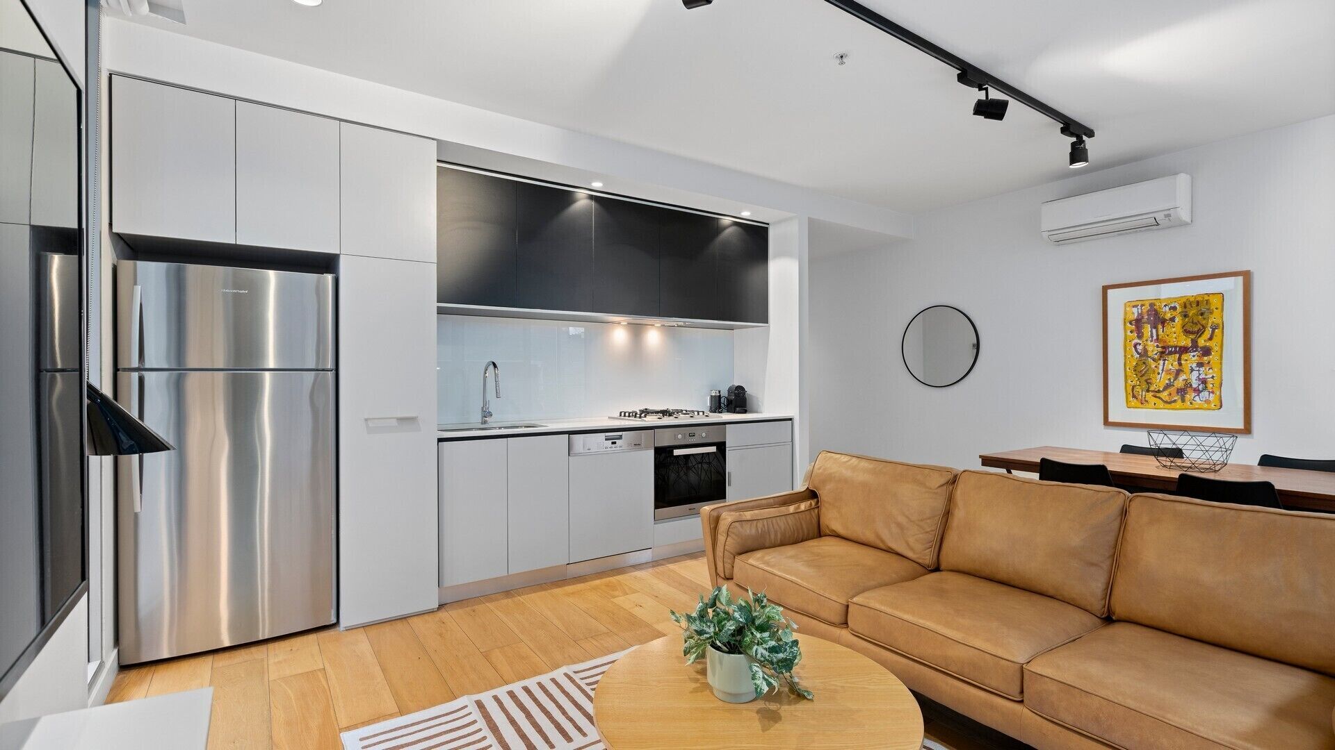 A modern kitchen with stainless steel appliances, a built-in oven, and a sink. There is a leather sofa and a round coffee table with a plant.