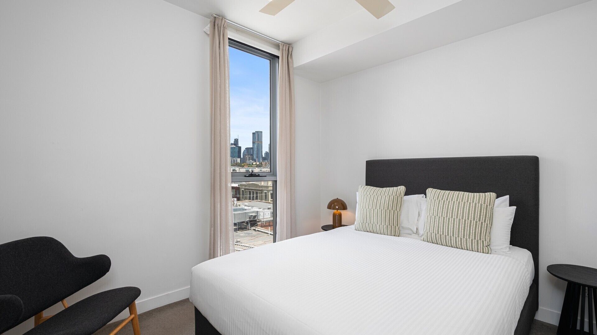 A modern hotel room with a large bed, a chair, a small table, and a view of the cityscape through a large window.