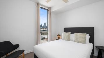 A modern hotel room with a large bed, a chair, a small table, and a view of the cityscape through a large window.
