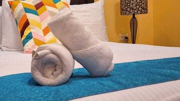 A neatly made bed with a blue blanket, a rolled white towel, and a multicolored pillow.