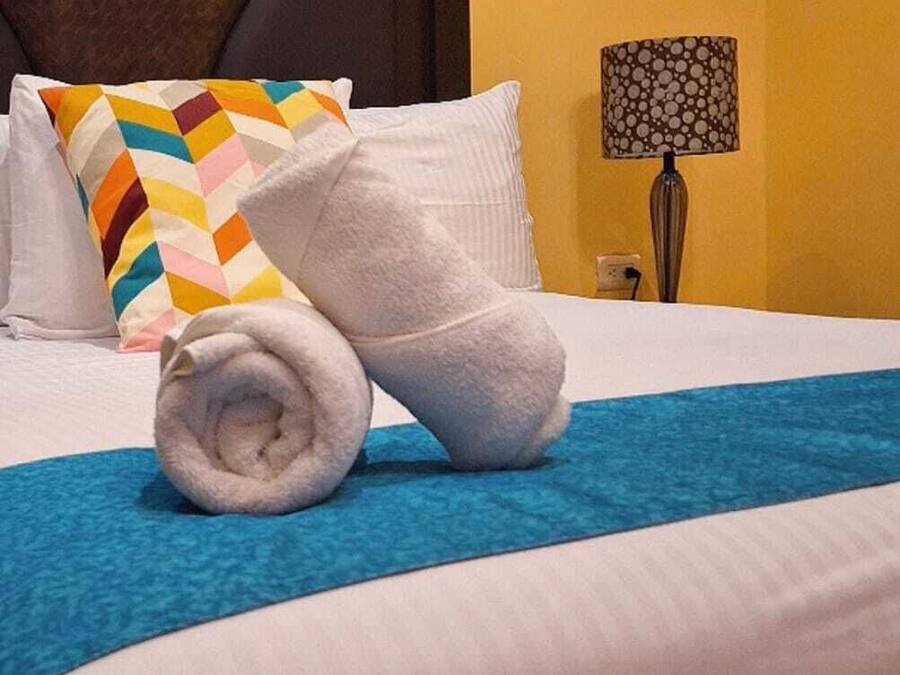 A neatly made bed with a blue blanket, a rolled white towel, and a multicolored pillow.